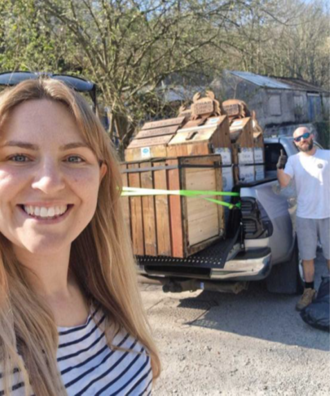 Large wooden crab line boxes attached to trailer in background. Smiling woman with long hair and stripey t-shirt in foreground. Man in white t-shirt and sunglasses to the right of the trailer.